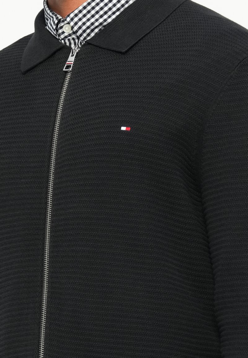 Black zip-up jacket featuring textured fabric, flat collar, and a small red, white, and blue logo. Interior shows checkered shirt collar.