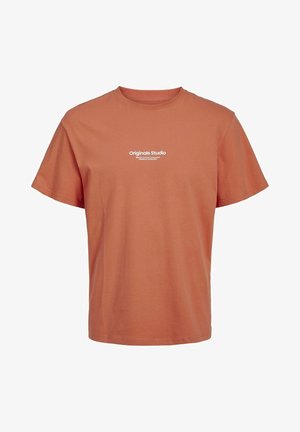 Homme portant un t-shirt orange « Originals Studio », un jean bleu clair et des baskets blanches avec des accents noirs, debout devant un fond uni.