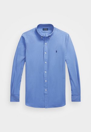 Camisa de manga larga azul claro con cuello abotonado, botones blancos y un pequeño logotipo bordado en el lado izquierdo del pecho. Textura suave.