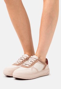 Cremefarbene und beige Sneakers mit strukturierten Wildlederakzenten und kontrastierendem roten Absatz. Sie verfügen über flache Schnürsenkel und eine dicke, gepolsterte Sohle.