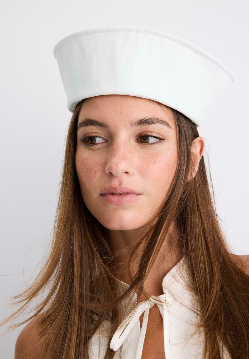 Jeune femme aux longs cheveux bruns portant un chapeau de marin blanc et un haut blanc avec un col noué, regardant légèrement sur le côté.