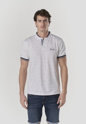 Polo blanca con patrón de puntos azules, cuello abotonado y puños con rayas azul marino. Se lleva con shorts de mezclilla y tiene un logo bordado.