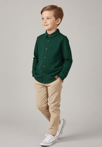 Zielona koszula z guzikami i kołnierzykiem, w połączeniu z beżowymi spodniami i białymi trampkami. Drewniane guziki i detal w postaci naszywki na koszuli.