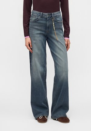 Person iført vidde-benet falmede blå jeans med en perlekæde som tilbehør på den forreste højre bæltestrop og brune sko.