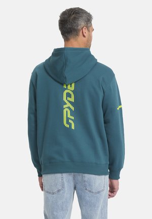 Ein Mann, der von hinten zu sehen ist, trägt ein türkisfarbenes Kapuzensweatshirt mit einem vertikalen gelben Motiv auf dem Rücken und hellblaue Jeans vor weißem Hintergrund.