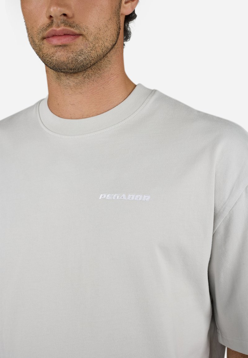 Helles graues T-Shirt mit kurzen Ärmeln aus Baumwolle, mit Rundhalsausschnitt und gesticktem „PEGADOR“-Logo in Weiß auf der Brust.