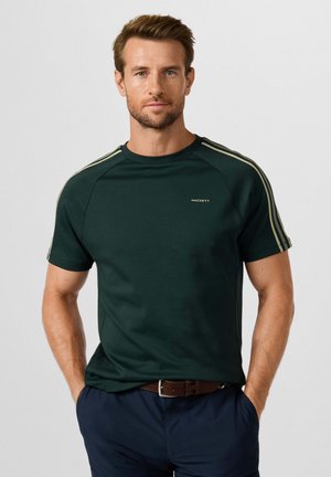 Hombre con pelo corto y castaño que lleva una camisa verde oscuro de manga corta con rayas beige en los hombros y pantalones azul marino.