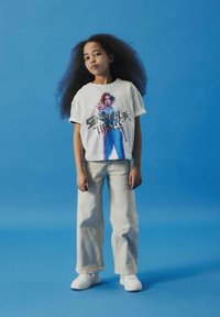 Hvid grafisk t-shirt med et farverigt "Stranger Things" design, parret med lysebeige vide bukser og hvide sneakers, mod en blå baggrund.