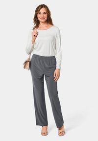 Grijze elastische broek gecombineerd met een lange mouwen witte top. Het model houdt een beige handtas vast en draagt een paar beige sandalen met bandjes. Strakke vormgeving.
