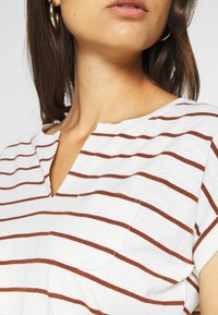 Haut en jersey blanc à manches courtes avec des rayures horizontales marron, doté d'un décolleté en V et d'une petite poche poitrine. Texture de tissu douce, design décontracté.