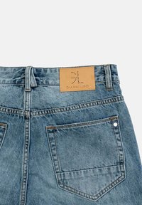 Jeans de mezclilla en azul claro con un parche de cuero etiquetado "GIANNILUPO". Presenta bolsillos traseros con costura visible y un pasador para el cinturón.