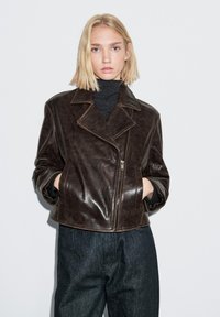 Braune Leder-Bikerjacke mit Reverskragen, Reißverschluss und Seitentaschen, kombiniert mit dunklen Jeans und einem schwarzen Rollkragenpullover.