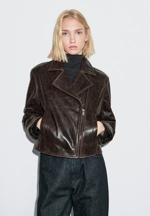 Braune Leder-Bikerjacke mit Reverskragen, Reißverschluss und Seitentaschen, kombiniert mit dunklen Jeans und einem schwarzen Rollkragenpullover.