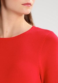 Punainen neulepaita, jossa on pyöreä kaula-aukko, sileä rakenne ja istuva muotoilu. Paidassa on saumaton viimeistely olkapäiden ympärillä.