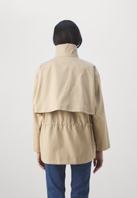 Chaqueta de algodón beige con un cuello alto, corte relajado y cintura ajustada. Presenta una solapa trasera sobredimensionada y mangas largas.