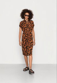Korte mouw oranje jurk met zwart luipaardprint, riem in de taille, knopen aan de voorkant en lengte tot de knie, gecombineerd met zwarte sandalen en een zonnebril.