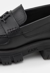 Des mocassins en cuir noir présentent un dessus texturé, des détails tissés et une semelle en caoutchouc épaisse avec le logo "KARL". Un design minimaliste avec des accents.