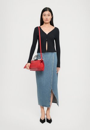 Femme portant un haut noir boutonné, une longue jupe en denim bleu avec fente à l'avant, des chaussures noires à bouts pointus et portant un sac à main rouge sur l'épaule.