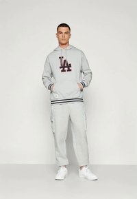 Jauns vīrietis stāv, ģērbies gaiši pelēkā kapucē ar "LA" logo, saskaņotās kravas sporta biksēs un baltās kedās, pret vienkāršu fona.