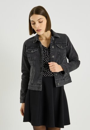 Femme aux cheveux bruns et lisses portant une veste en denim noire sur un chemisier noir à pois et une jupe noire, regardant vers le bas avec une main posée sur la veste.
