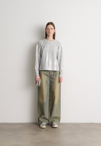 Mulher encostada a uma parede branca, vestindo um suéter de lantejoulas prateadas, calças largas desbotadas, ténis prateados, e segurando uma pequena clutch prateada.