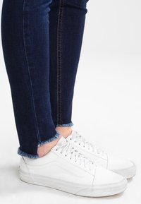 Person, der abgerissene, dunkelblaue Skinny Jeans mit fransigem Saum und weiße, flache Schnürsneaker auf einem weißen Hintergrund trägt.