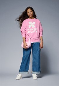 Roze sweatshirt met witte grafische tekst, gecombineerd met wijde denim jeans opgerold bij de manchetten en witte sneakers. Draagt een roze tas.
