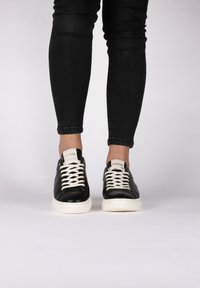 Černé kožené tenisky s bílými tkaničkami a bílou gumovou podrážkou. Obsahují logo na jazyku a očka na tkaničky. Hladký povrch.