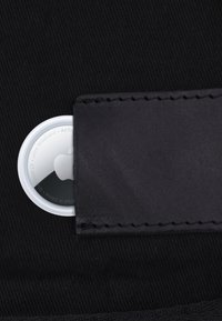 Apple AirTag częściowo włożony do czarnego skórzanego portfela na czarnym materiałowym tle.
