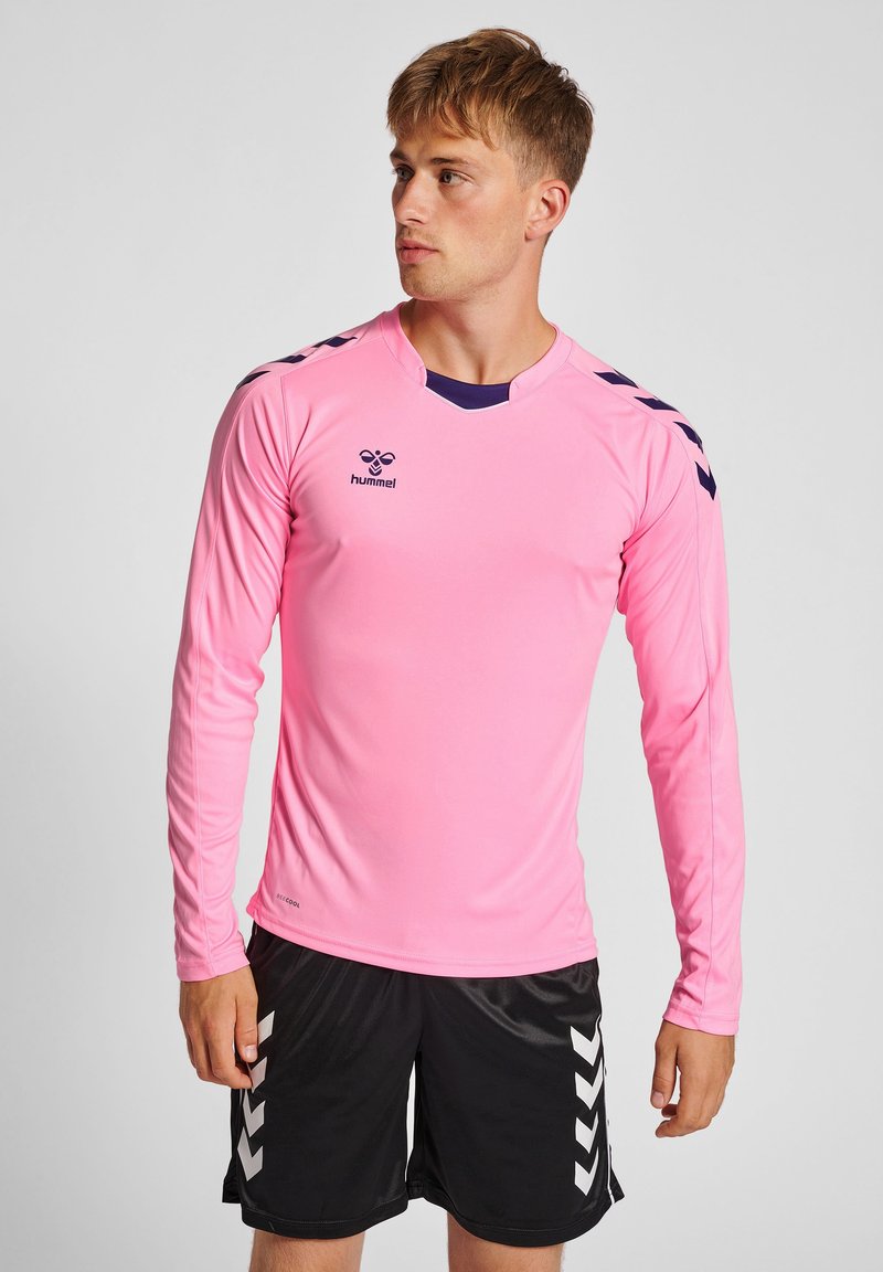 Maglietta sportiva a maniche lunghe di colore rosa con dettagli blu navy, caratterizzata da un collo a V e un logo. Abbinata a pantaloni corti neri con strisce bianche a zigzag.