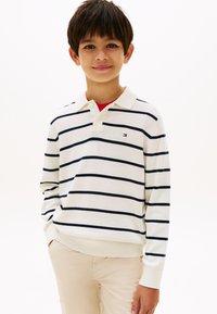 Αγόρι με σκούρα μαλλιά που φοράει λευκό πόλο μακρυμάνικο με μαύρες ρίγες και μπεζ παντελόνι, στέκεται με το ένα χέρι στην τσέπη, μπροστά σε λευκό φόντο.