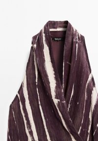 Vêtement sans manches violet et crème avec un col châle, présentant un tissu texturé avec de larges rayures verticales. Étiquette de marque visible.