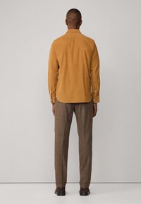 Camisa de corduroy marrón claro con cuello abotonado, mangas largas y bolsillo frontal. Combinada con pantalones texturizados marrones y zapatos oscuros.