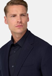 Marinblå herringbone-blazer med spetsig krage, en bröstficka och mörk skjorta, med mjukt tyg och skräddarsydd passform.