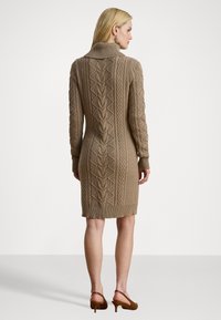 Blond kvinna klädd i en knälång brun kabelstickad klänning med hög krage, tillsammans med bruna klackslippers, vänd bort.