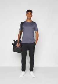 Grå polyester treningst-skjorte, svarte bomullssweatpants, hvite joggesko og en svart skulderveske med glidelås, båret av en stående figur.