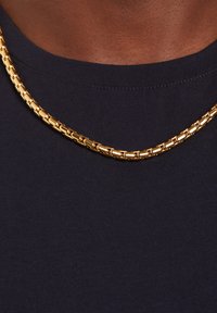 Gouden ketting met schakels die in elkaar grijpen, met een glanzende afwerking. Draagt over een donkerblauw shirt, waardoor de metaalachtige textuur wordt benadrukt.