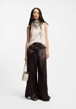 Frau trägt ärmellose cremefarbene Bluse, leicht gemusterten Schal, weite schwarze Satin-Hosen, schwarze Sandalen, hält eine beige Handtasche und steht vor weißem Hintergrund.