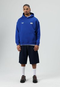 Blauwe hoodie met een kangoeroezak, witte accenten en logo; gedragen met donkere denimshorts en zwarte sneakers. Witte sokken zichtbaar.