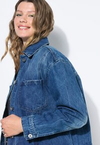 Jeansjacka i medelblått med en klassisk krage, knäppning, framficka och uppkavlade ärmar. Har en något sliten textur.