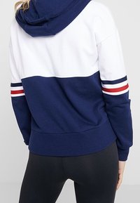 Sweatshirt à capuche bleu marine et blanc avec des rayures rouges et blanches sur les manches. Présente une coupe décontractée ainsi que des poignets et un ourlet en rib. 