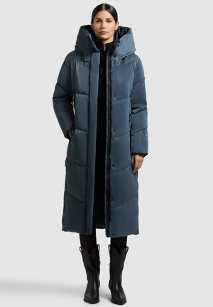 Abrigo acolchado largo en un tono verde azulado oscuro con capucha, que presenta un cierre de cremallera y botones a presión, paneles texturizados y bolsillos laterales. Llevado sobre un atuendo negro.