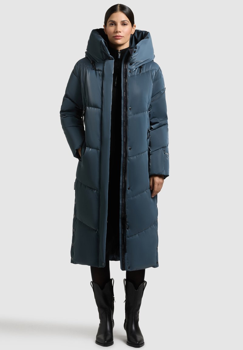 Lang, donkerblauw-groene pufferjas met een capuchon, voorzien van een rits en drukknoopsluiting, textuurpanelen en zijzakken. Draagt over zwarte kleding.