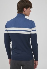 Blauer und marineblauer Pullover mit hohem Kragen, der ein weißes und dunkelblaues Streifenmuster über dem oberen Rücken und an den Ärmeln aufweist.