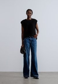 Fekete ujjatlan kötött felső, bő szárú kék farmernadrág és egy kis fekete kézitáska, egyszerű háttérrel és semleges világítással.