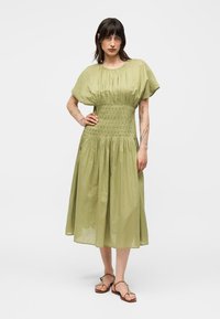 Naiste tume juustega, kandes heledat rohelist lühikeste varrukatega keskjoonega midi-kleiti ja pruune sandaale, seisab ühe käega puusal.