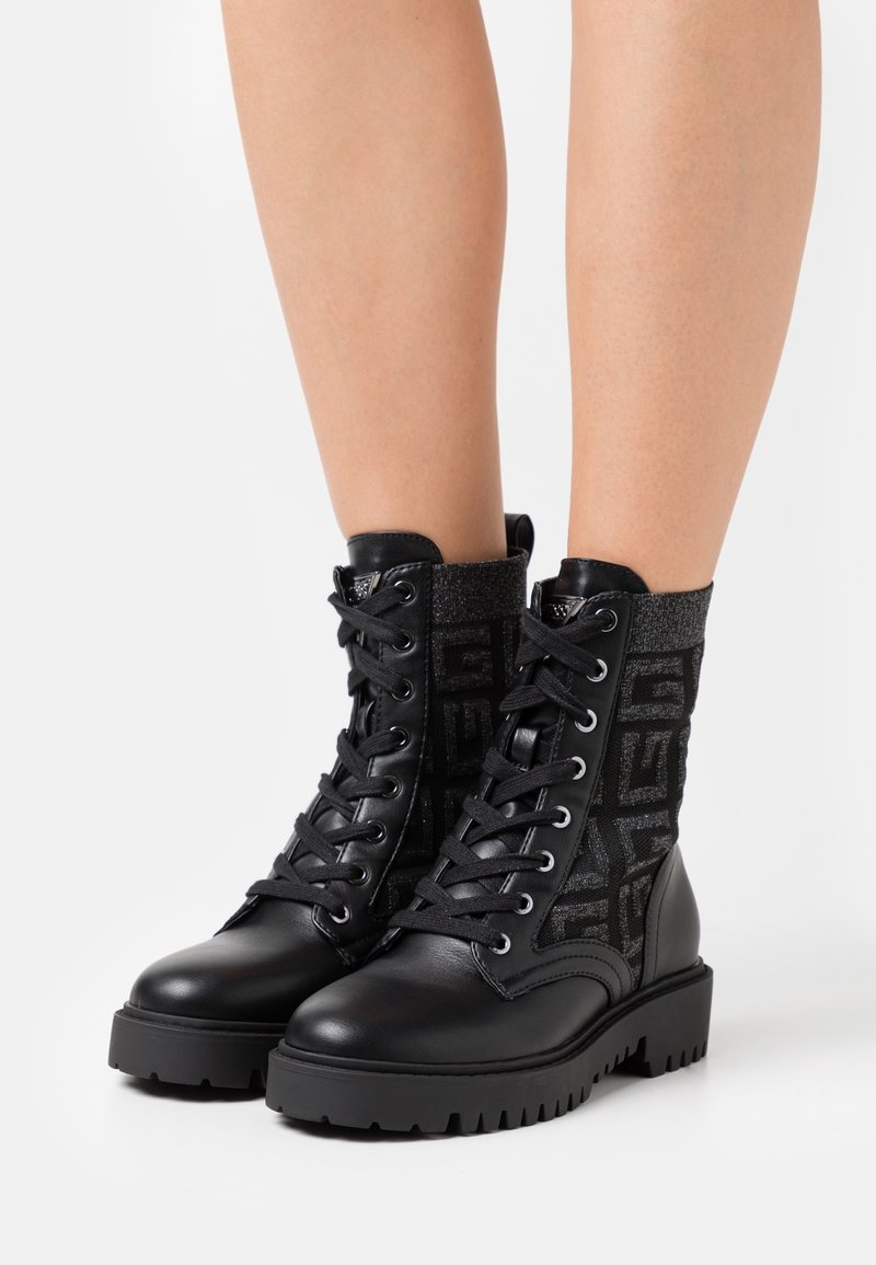 Bottes de combat noires avec un panneau en tissu texturé à motif, corps de chaussure en cuir, bout rond et semelle épaisse en caoutchouc.