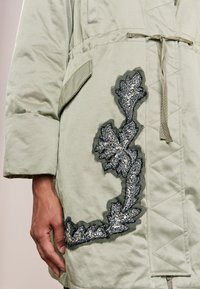 Giacca esterna imbottita verde con ricamo di foglie testurizzato e dettagli metallici, vita regolabile e tasche inclinate.