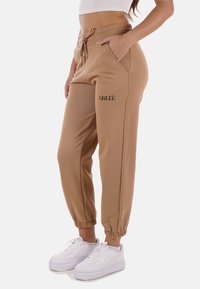 Pantaloni jogger khaki con polsini elasticizzati, vita con cordoncino, tasche laterali e testo stampato nero sulla coscia. Realizzati in tessuto morbido.