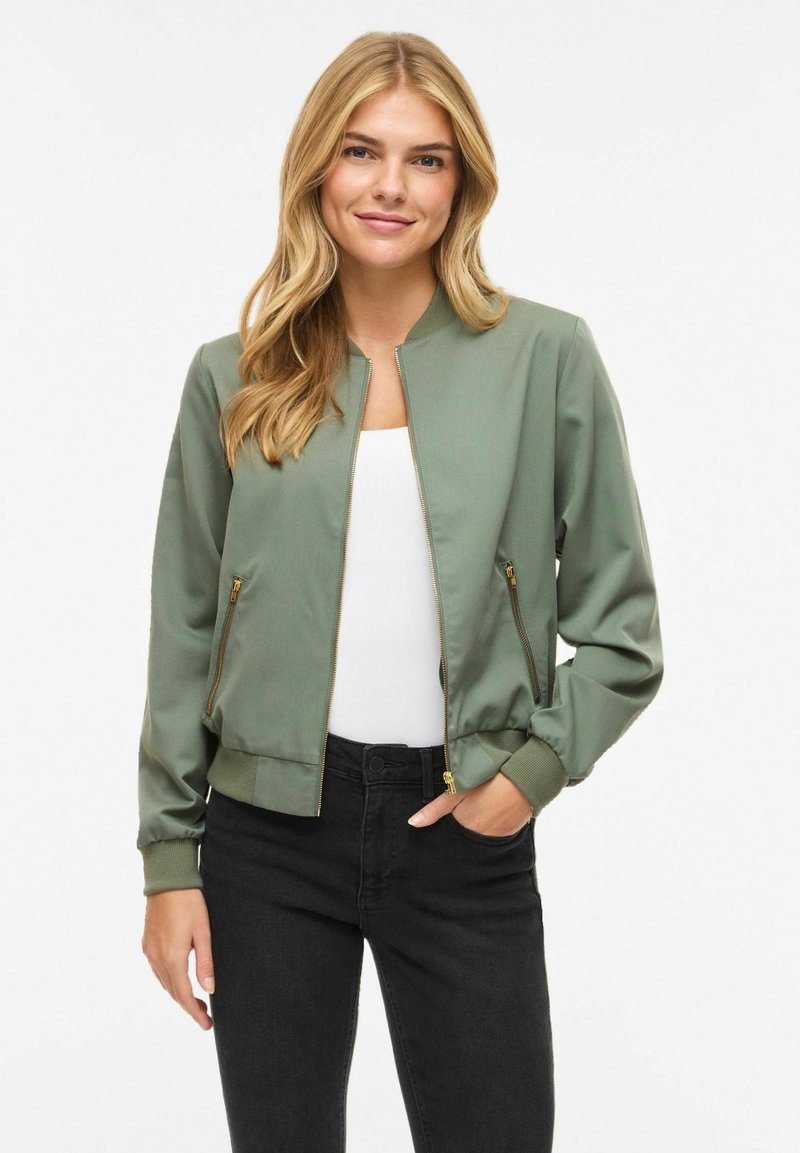 Veste bomber vert olive, en tissu lisse. Comprend une fermeture éclair, des poignets côtelés et des poches latérales avec des accents dorés.